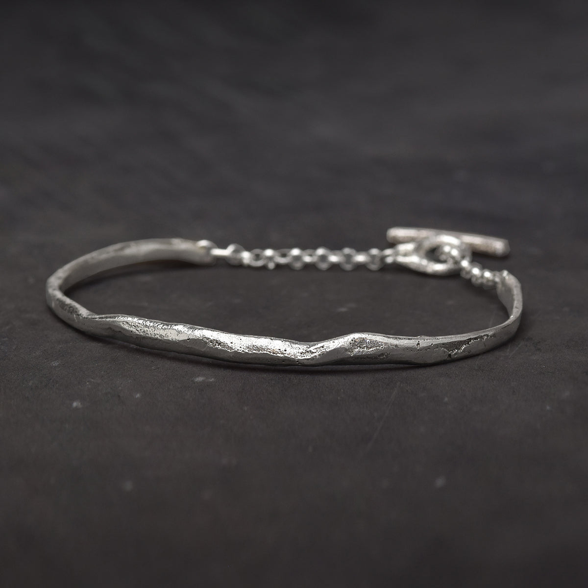 Slightly textured sterling silver cuff bracelet with a melted shape and a chain with toggle clasp on a dark background.