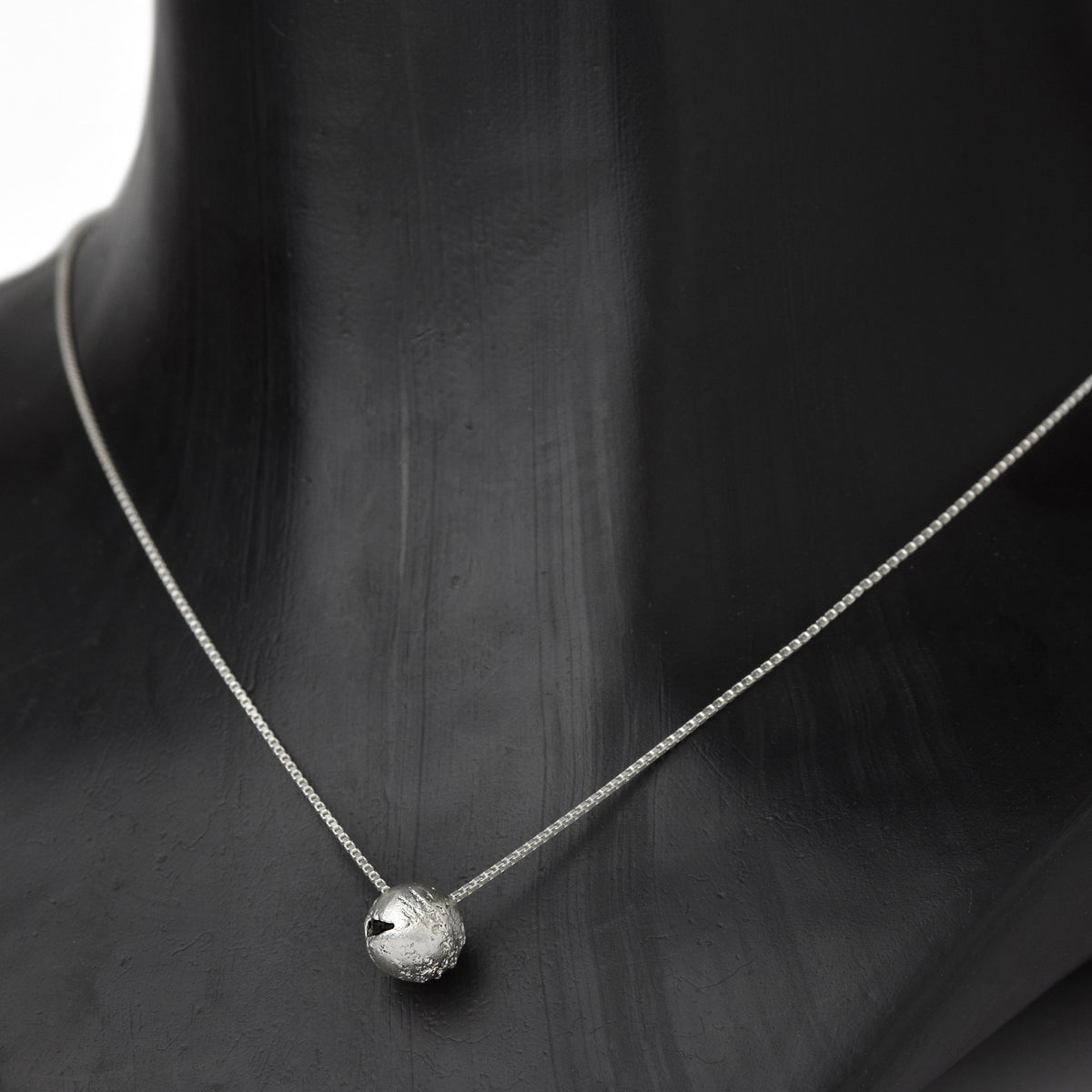 Handcrafted sterling silver hollow sphere pendant with a textured finish suspended from a sterling silver chain on a dark neck.